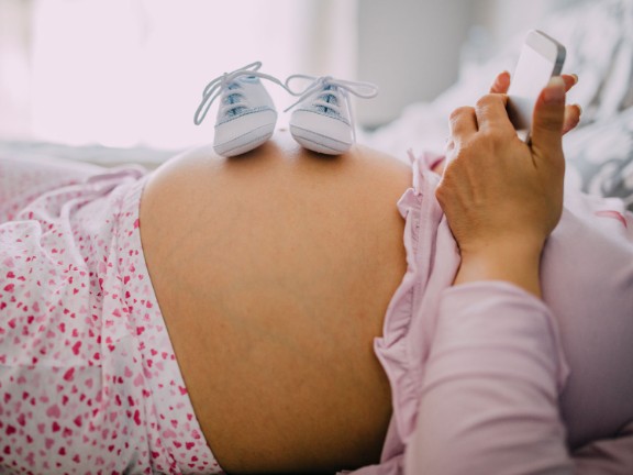 Woodland Memorial Hospital Family Birth Center | Dignity Health Pregnant woman with baby shoes on her tummy and looking at cell phone.